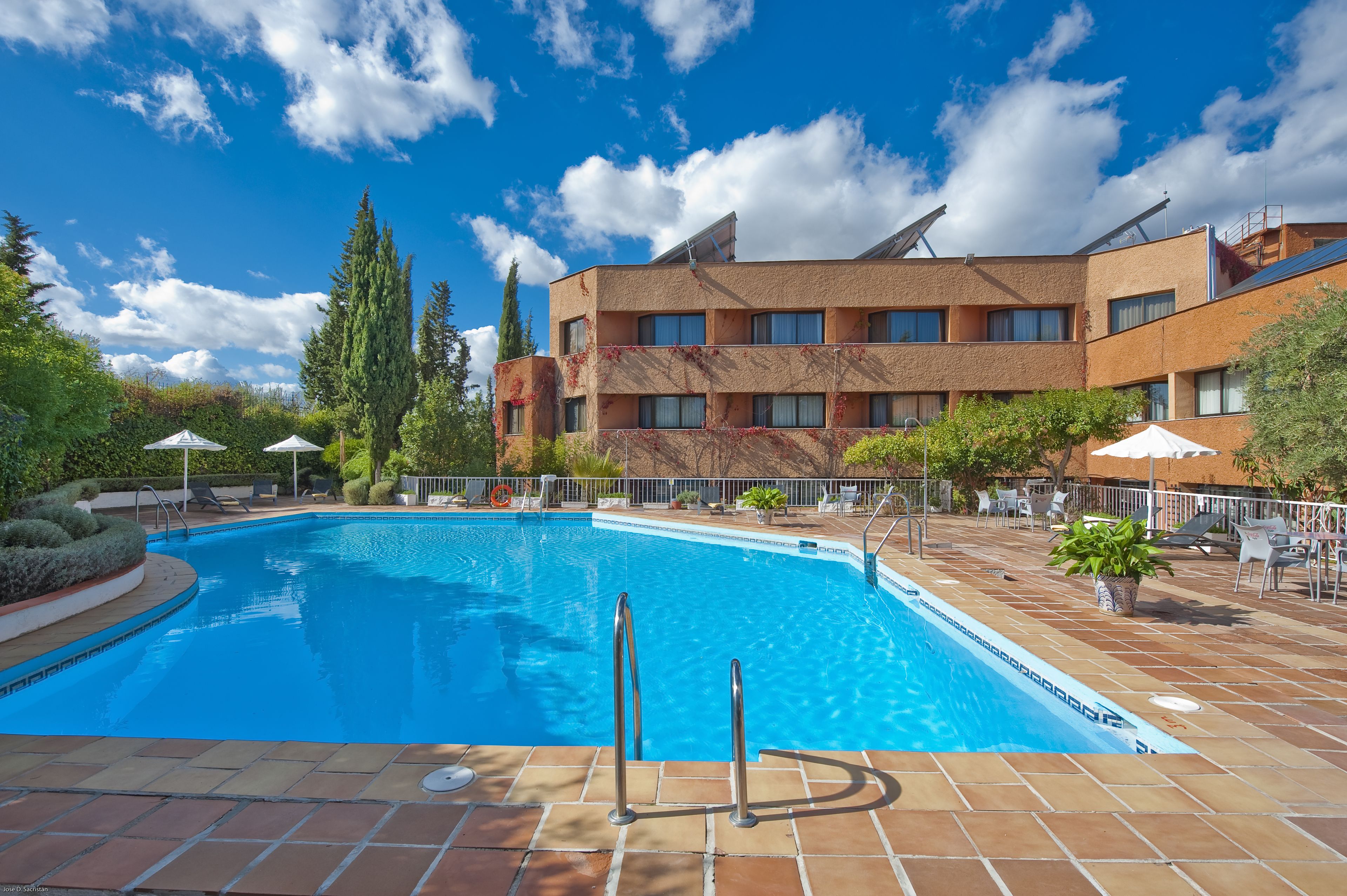 A swimming pool with a tiled deck, surrounded by lounge chairs and umbrellas, and a building with a modern design in the background.