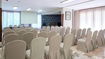 Un salón de conferencias con filas de sillas color crema, una pantalla de proyector y un amplio ventanal con cortinas.