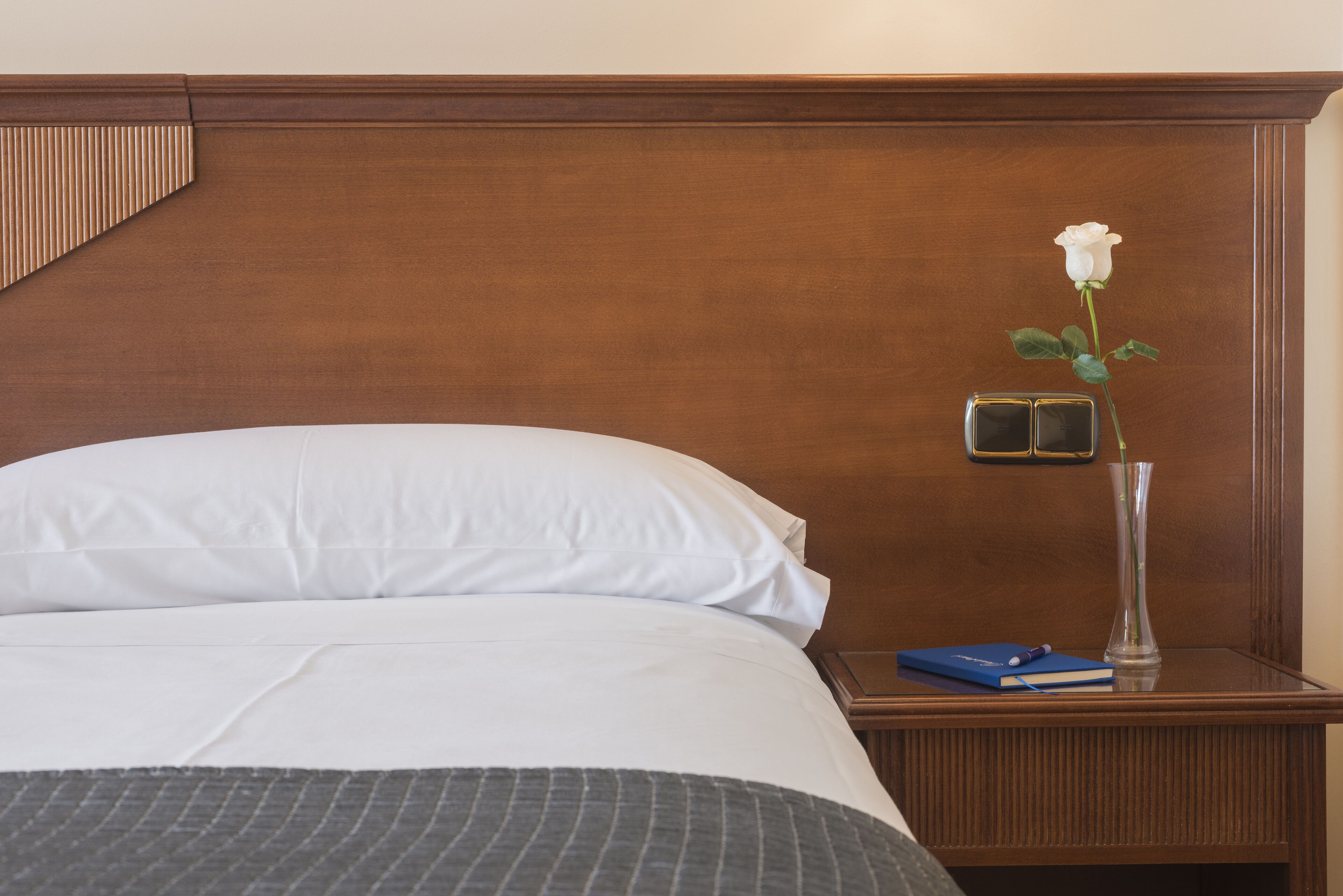 A neatly made bed with white linens, a bedside table with a vase of a white rose, and a blue book.