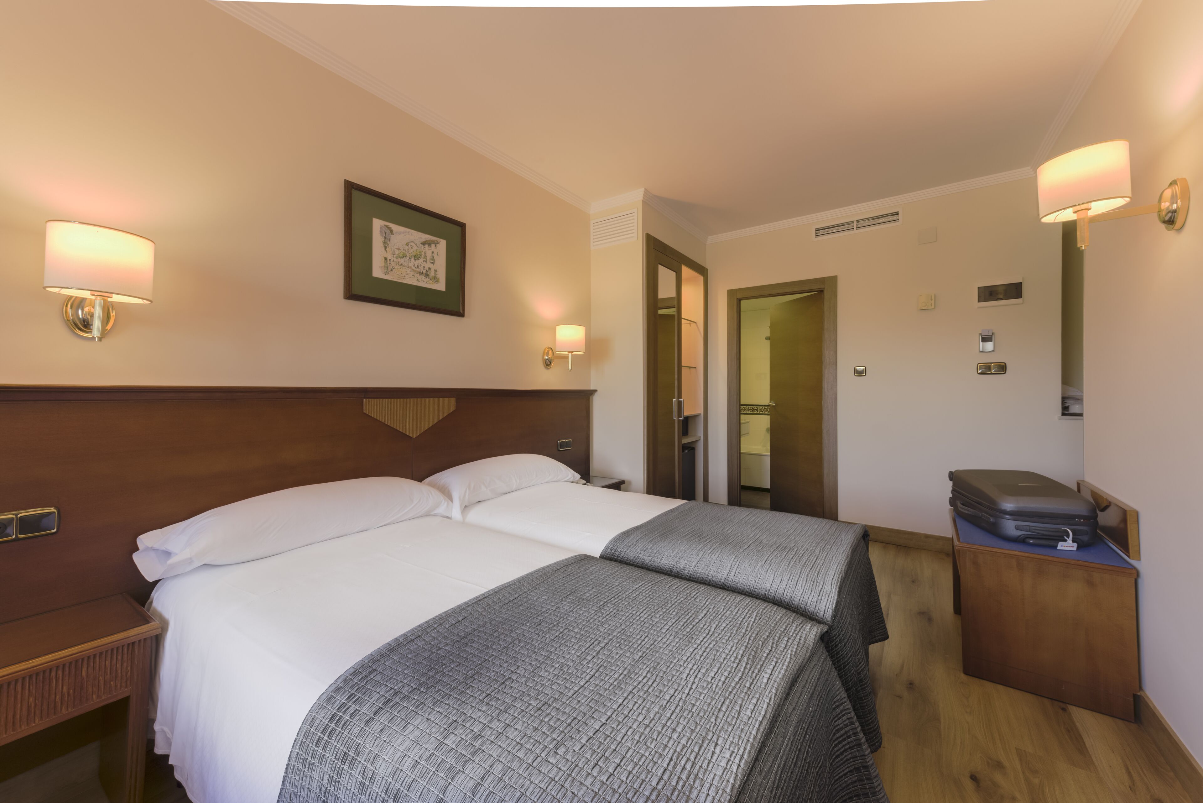 Habitación de hotel con una cama doble, dos mesitas de noche, un cabecero de madera, un escritorio pequeño con una maleta y lámparas fijadas a la pared.