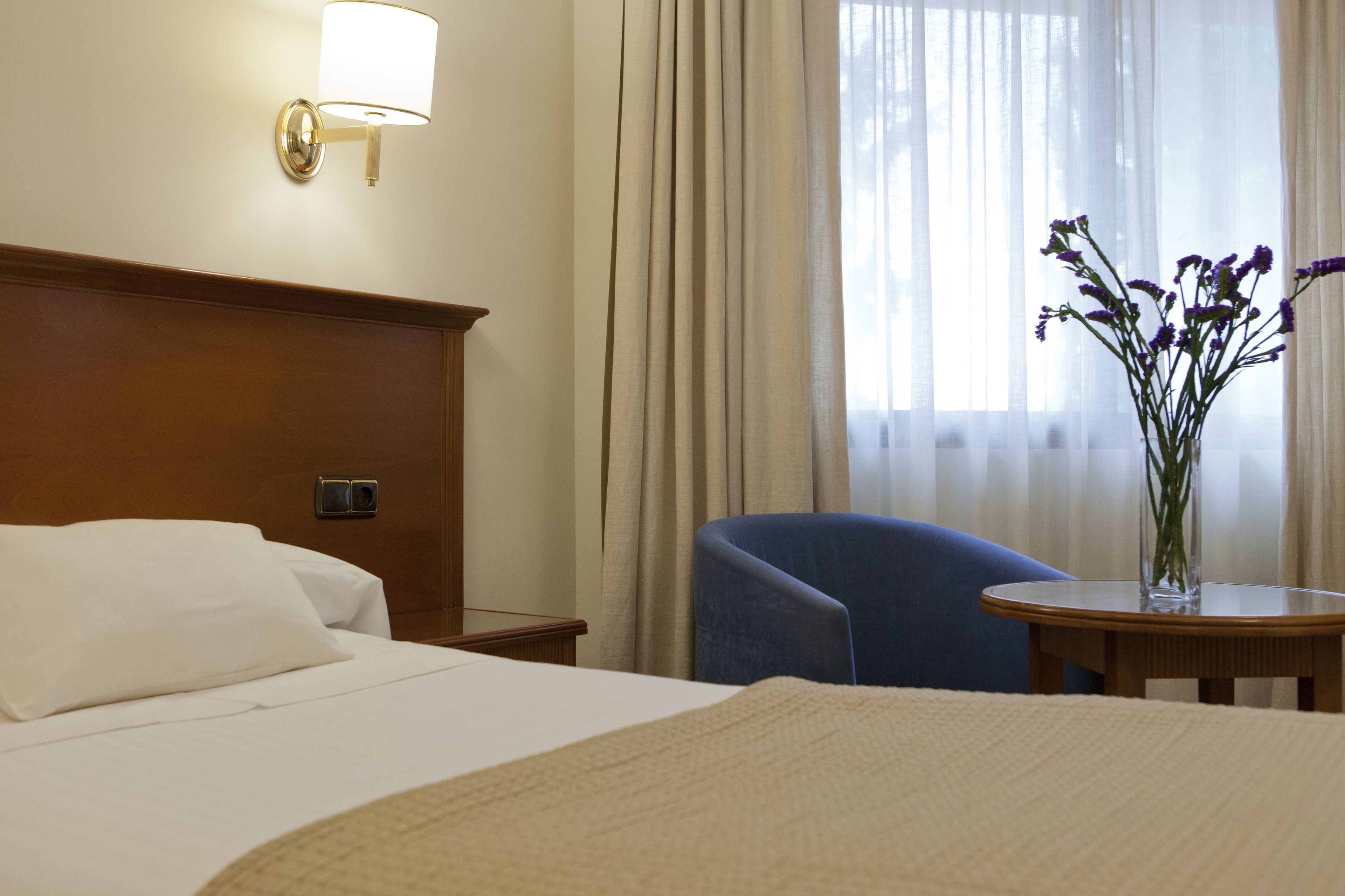 A hotel room with a bed, a nightstand, a lamp, a chair, a table, and a vase with flowers.