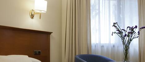 A hotel room with a bed, a nightstand, a lamp, a chair, a table, and a vase with flowers.