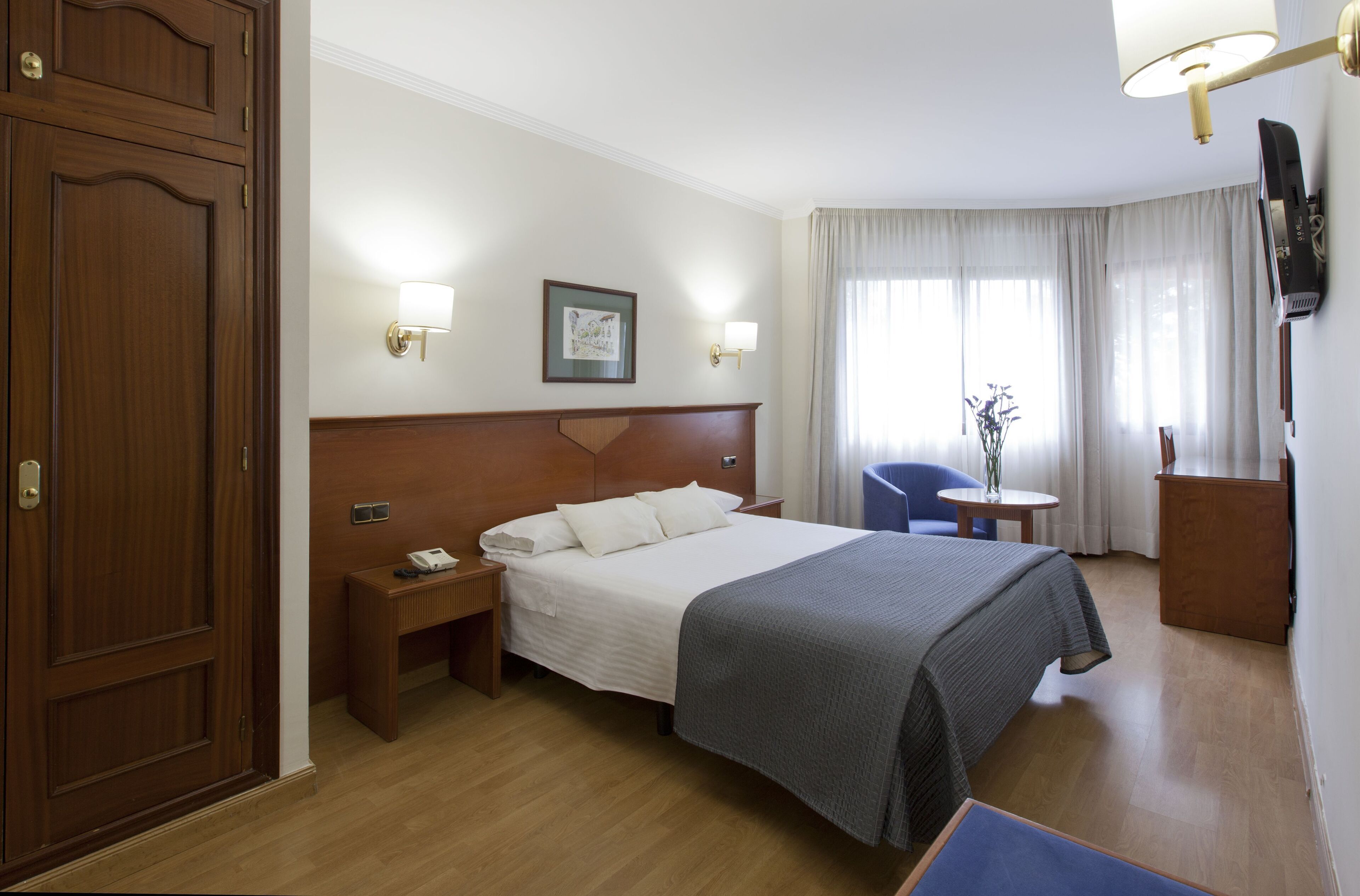 Una habitació d'hotel amb llit de fusta, taules de nit, escriptori, cadira i televisió penjada a la paret.