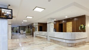 Et hotellets reception område med marmorgulv, en receptionsdisk og et fjernsyn, der viser hotelværelser.