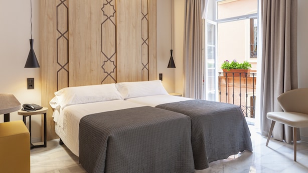 A double bed with a white and gray bedspread, a bedside table with a lamp, a small ottoman, and a view of the outside balcony with plants.