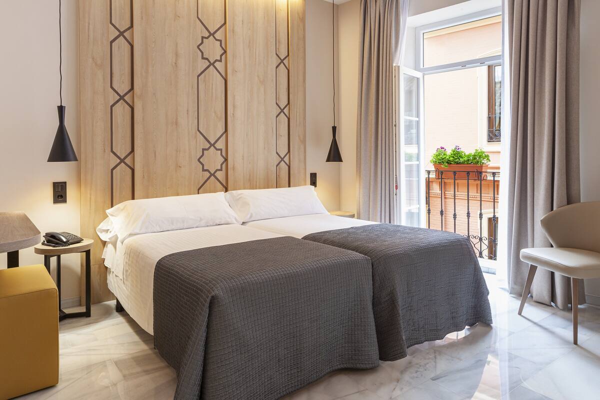 A double bed with a white and gray bedspread, a bedside table with a lamp, a small ottoman, and a view of the outside balcony with plants.