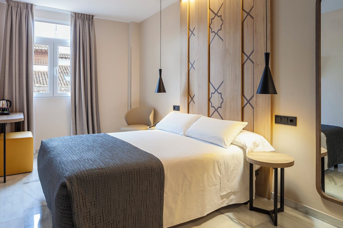 A hotel room with a bed, bedside table, chair, and a window with curtains.