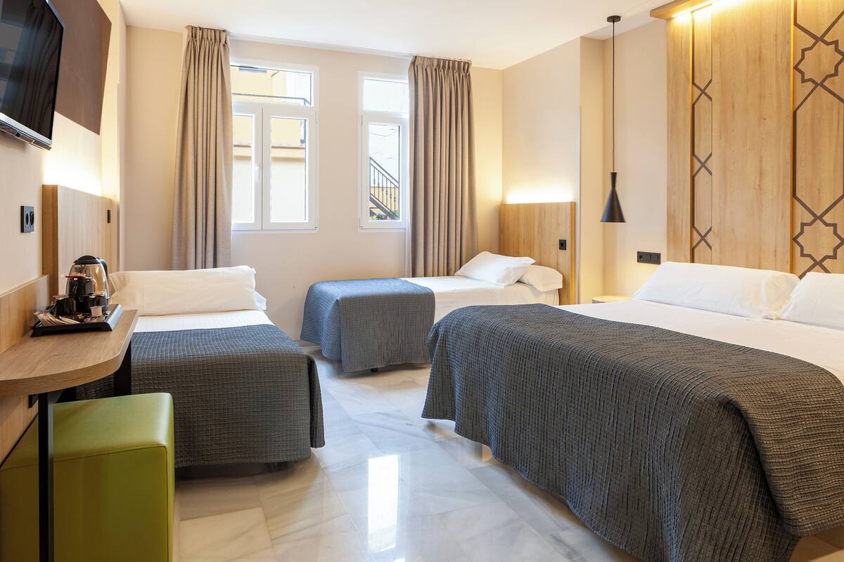 A hotel room with three beds, a small table with a kettle, a green chair, and a TV mounted on the wall.