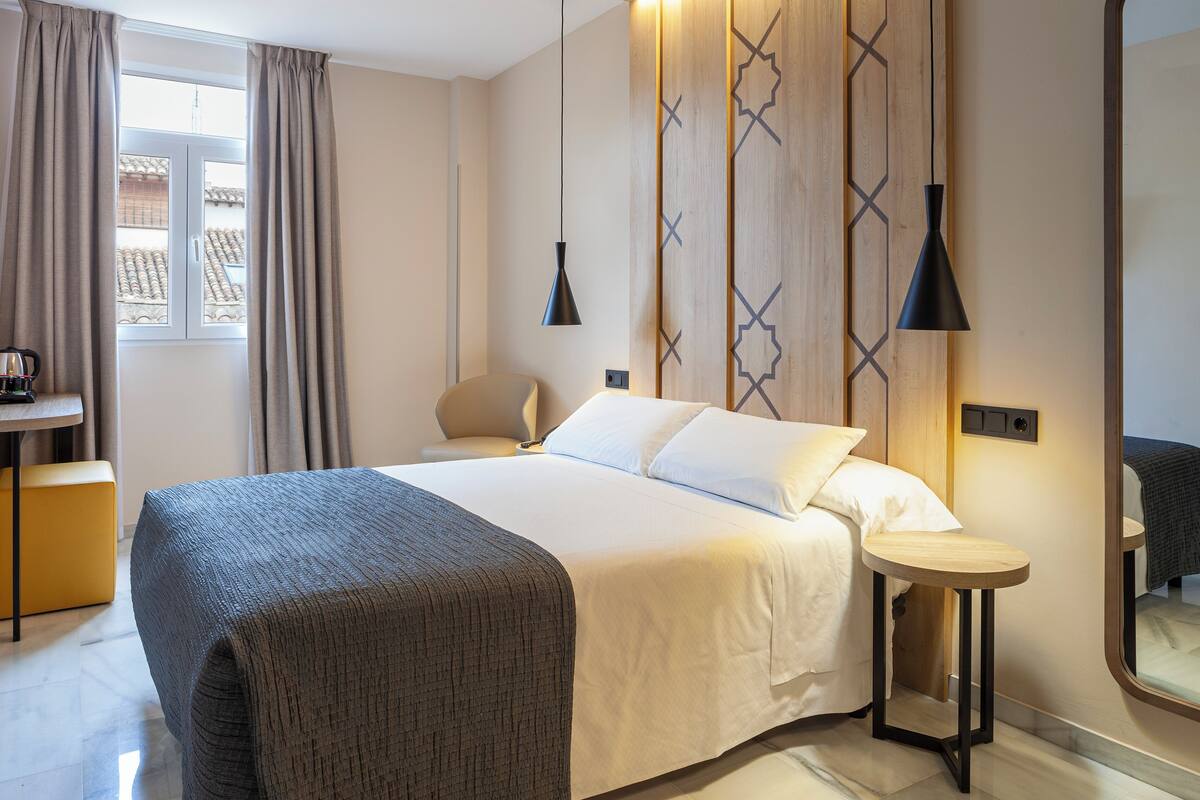 A hotel room with a bed, bedside table, chair, and a window with curtains.
