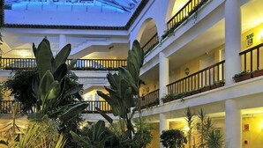 Un atrio de varios niveles con techo de vidrio, múltiples balcones y un patio central con suelo a cuadros y plantas en macetas.