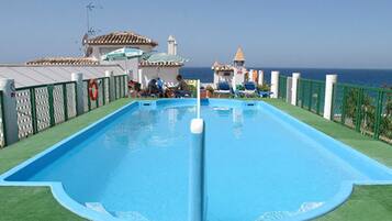 Una piscina rectangular con un flotador salvavidas, rodeada por una valla verde, y un edificio con techo de tejas al fondo.
