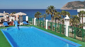 Una piscina rectangular con agua azul cristalina, rodeada de césped artificial verde y con vistas al mar y una colina al fondo.
