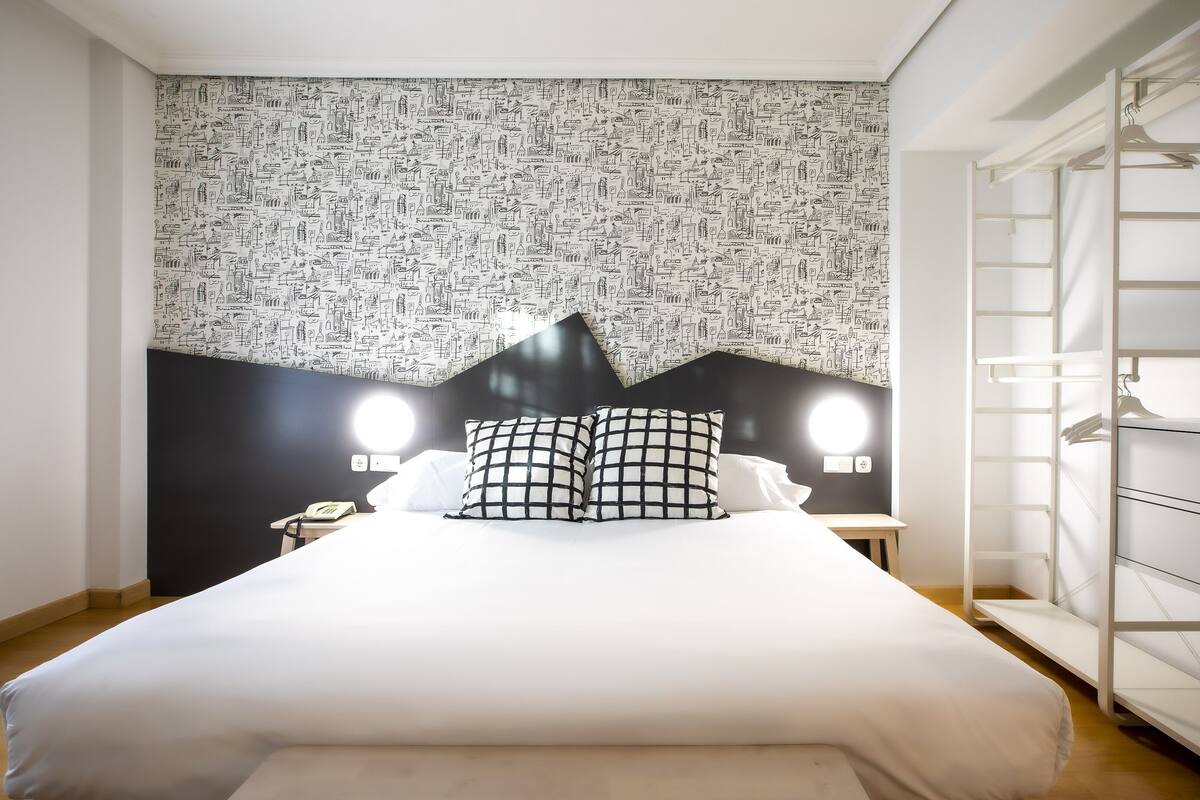 Uma cama bem arrumada com travesseiro quadriculado preto e branco, cabeceira preta e parede com padrão de diversos ícones.