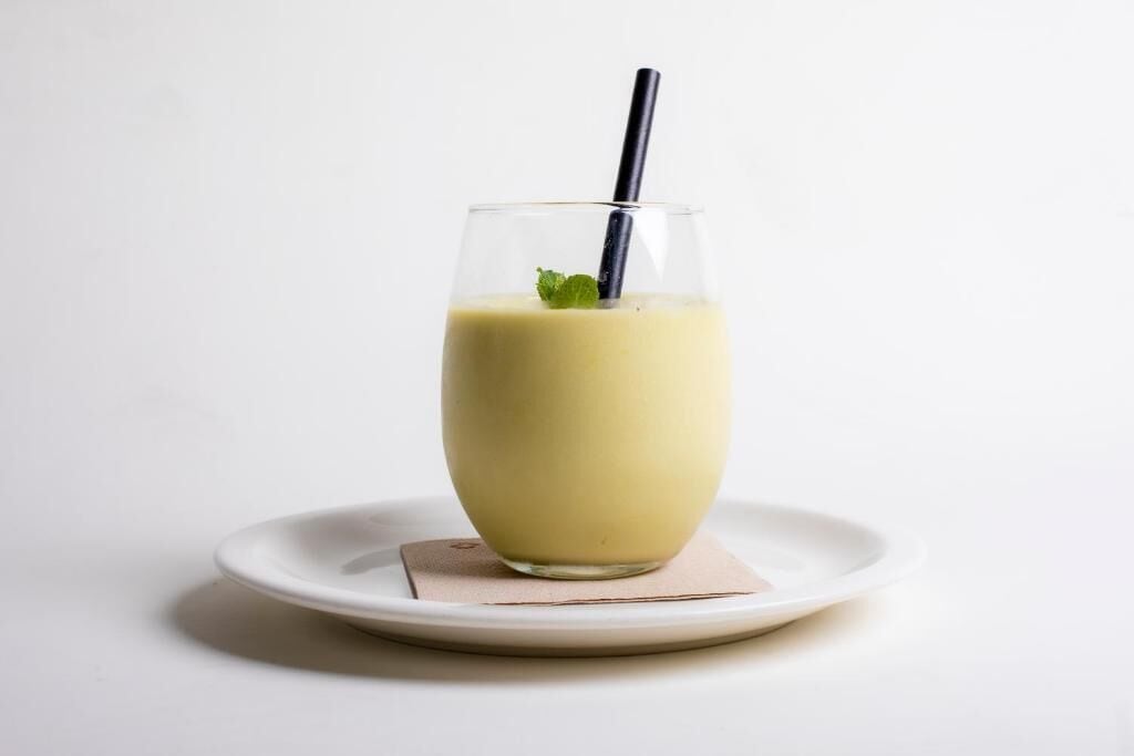 A glass of yellow beverage on a white plate with a black straw and a mint leaf garnish.