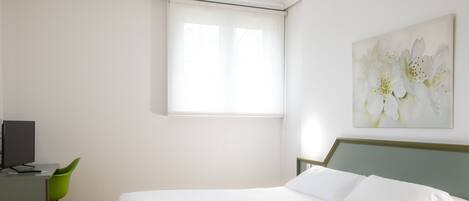 A bedroom with a bed, a desk with a computer, a green chair, and a window with blinds.
