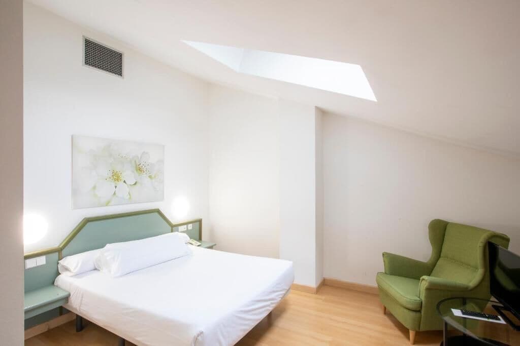 A bedroom with a bed, a green armchair, a desk with a computer, and a skylight.