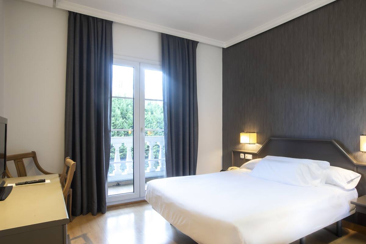 Quarto de hotel com cama, escrivaninha, cadeira, mesa de cabeceira e vista para o verde pela janela.