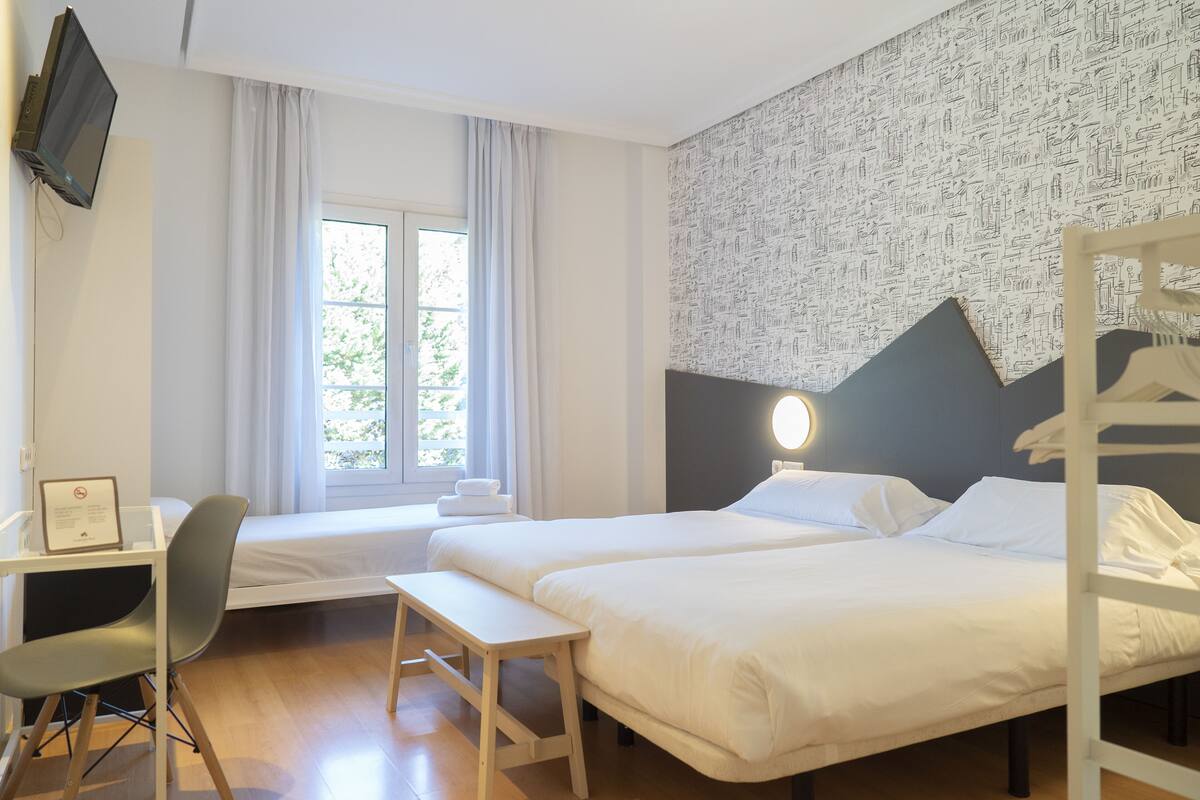Quarto de hotel com duas camas, uma mesa, uma cadeira e uma janela com cortinas.
