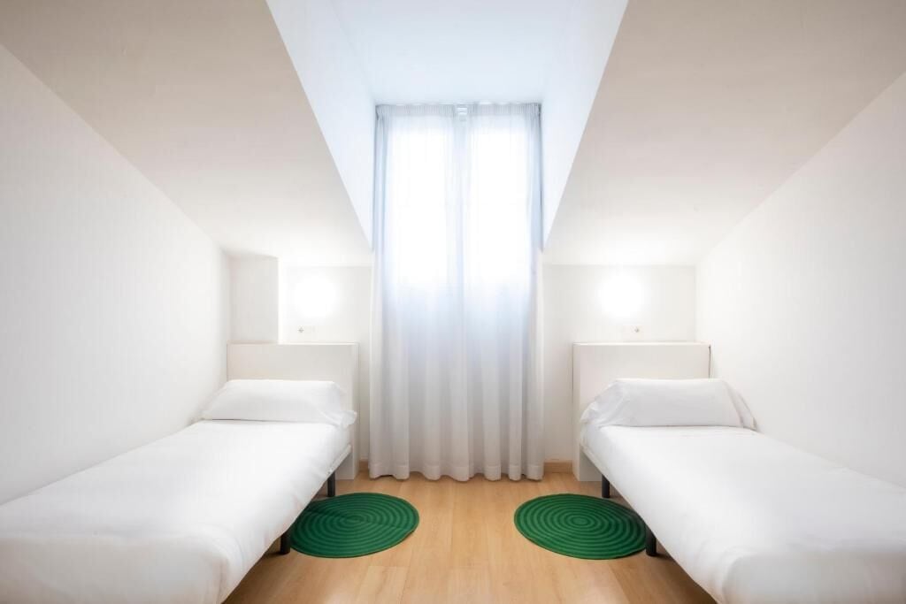 Une petite chambre moderne avec deux lits simples, une grande fenêtre avec des rideaux légers et un plancher en bois.