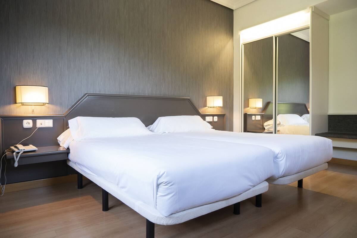 Quarto de hotel moderno com uma cama grande, luminárias laterais, um telefone na mesinha de cabeceira e um espelho na parede.