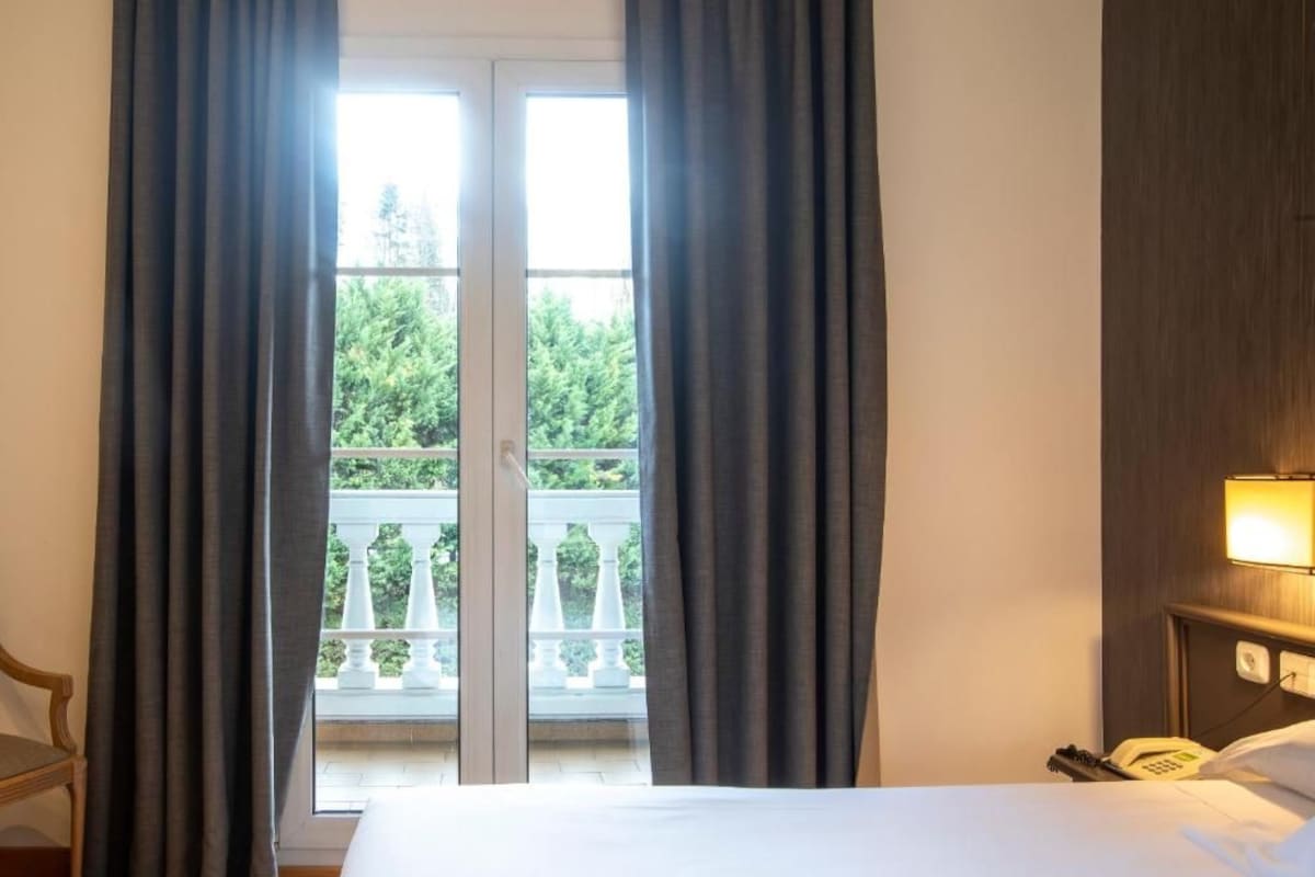 A hotel room with a bed, a nightstand with a phone, and a balcony with white railings and greenery outside.