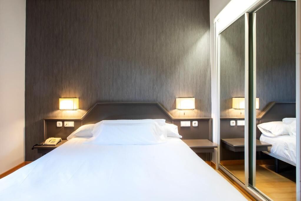 Una habitación de hotel con una cama grande, dos mesitas de noche, un espejo y lámparas de pared.