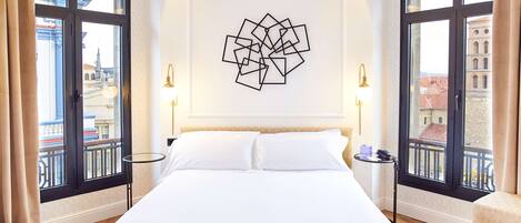 A hotel room with a large bed, two bedside tables, a wall art piece, and large windows with a city view.