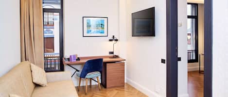 Una habitación de hotel moderna con piso de madera, un sofá color beige, un escritorio con silla, un televisor montado en la pared y un cuadro enmarcado.