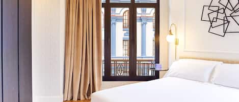 A hotel room with a bed, a window with curtains, a nightstand with a lamp, and a wall with a geometric art piece.