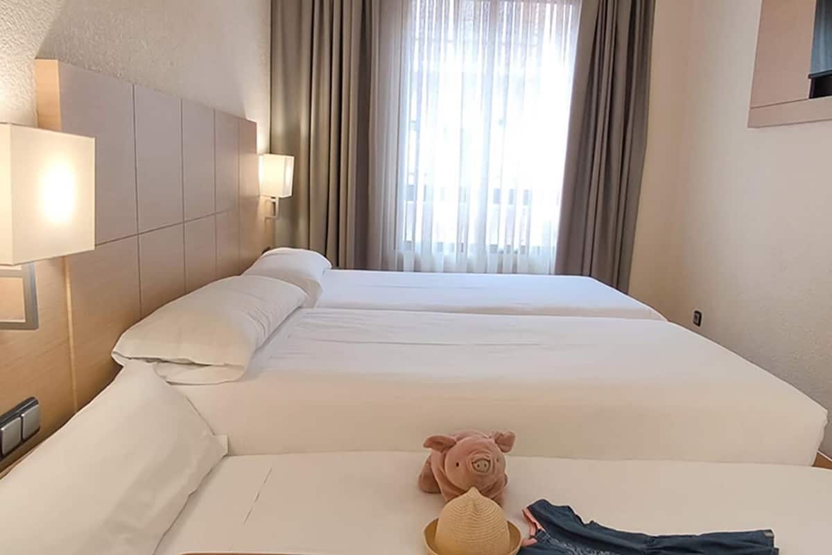 A hotel room with two beds, a nightstand, a lamp, a window with curtains, and a pig-shaped pillow.