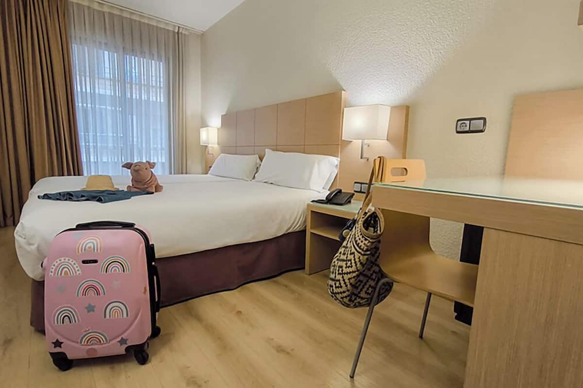 A hotel room with a bed, a desk, a lamp, a phone, a suitcase, and a handbag.