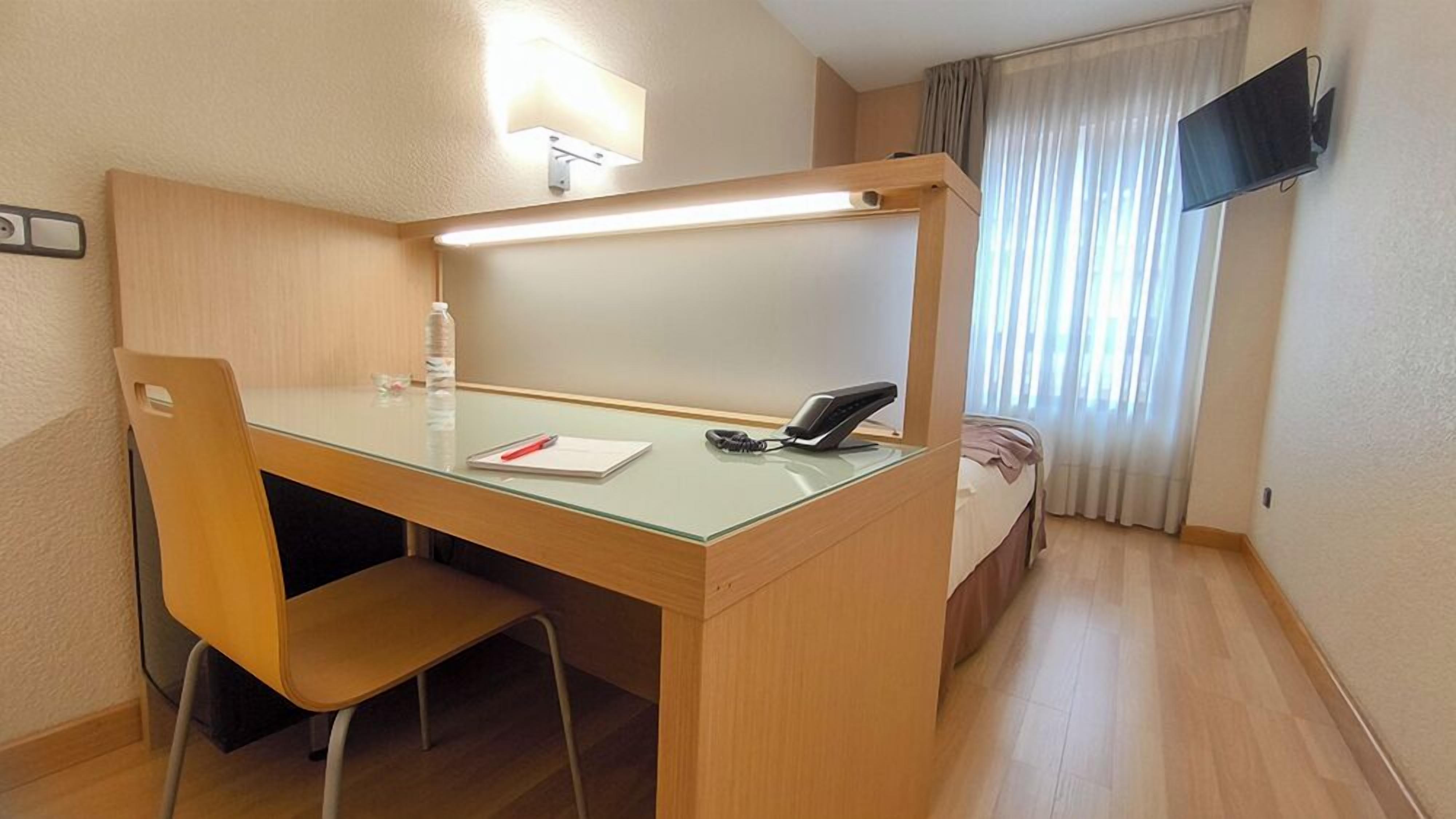 Habitación de hotel con escritorio, silla, cama, mesita de noche, lámpara y televisor montado en la pared.