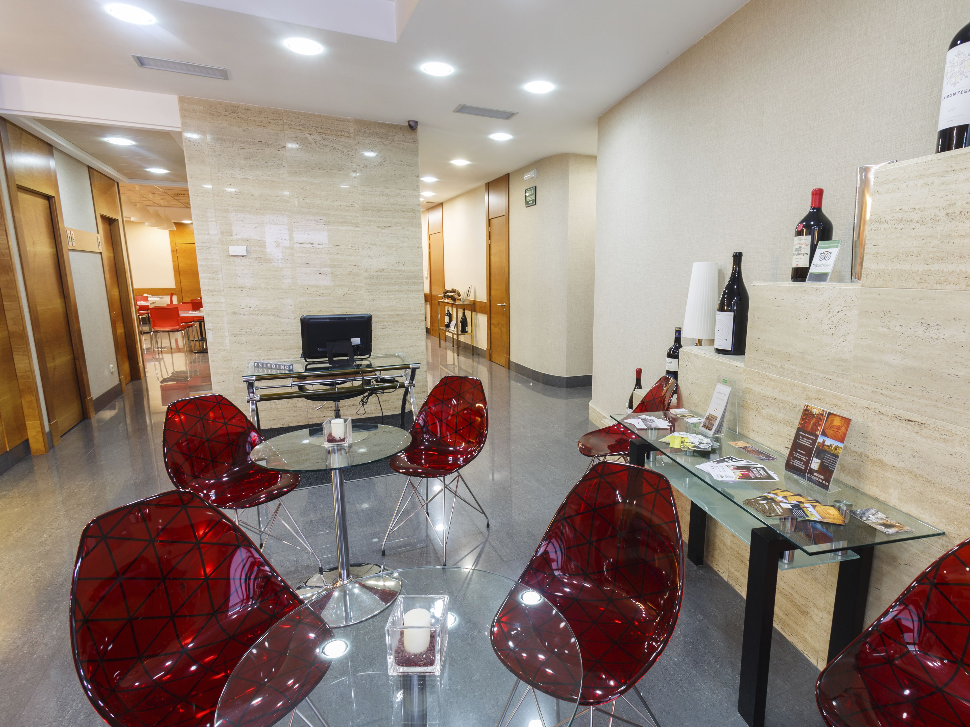 Un área de comedor moderna con mesas de vidrio y sillas rojas, un televisor y botellas de vino en una estantería.