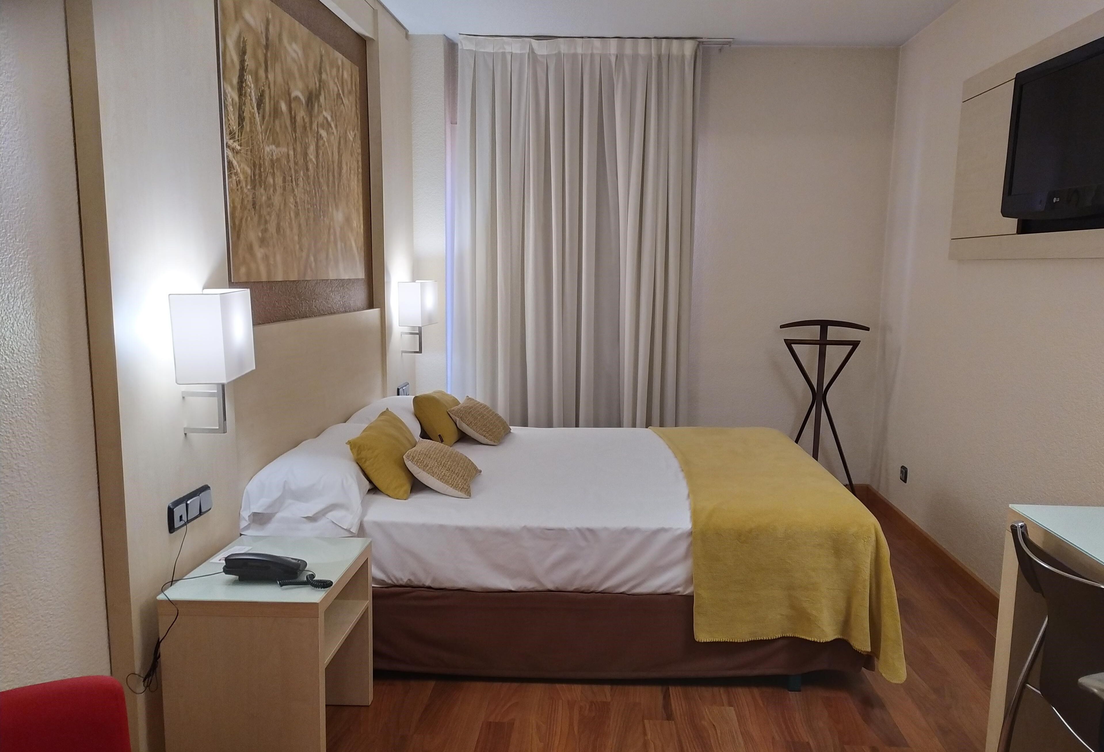 Una habitación de hotel con cama, mesita de noche, lámpara y televisor montado en la pared.