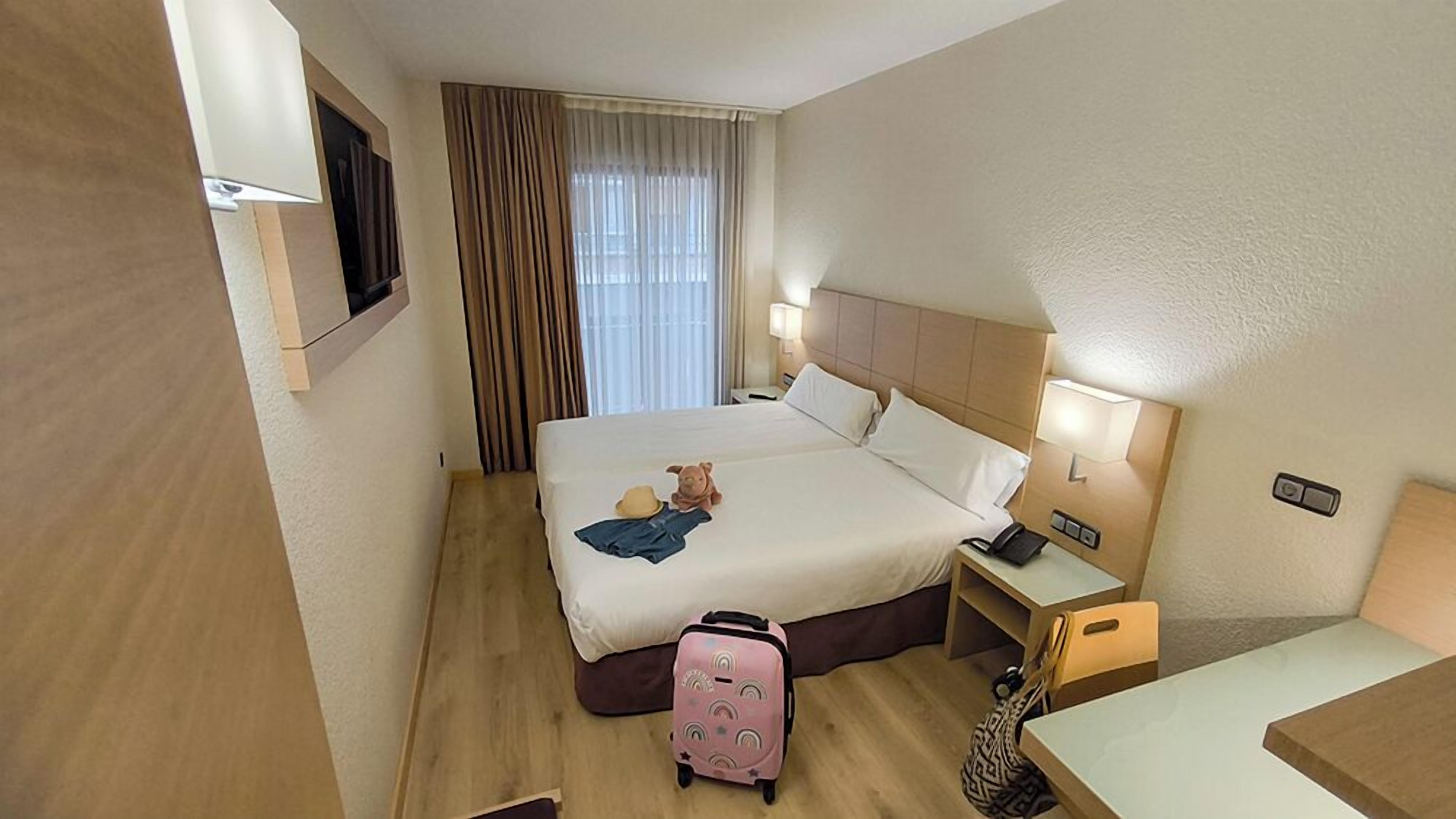 A hotel room with a bed, bedside tables, a lamp, a suitcase, and a TV mounted on the wall.