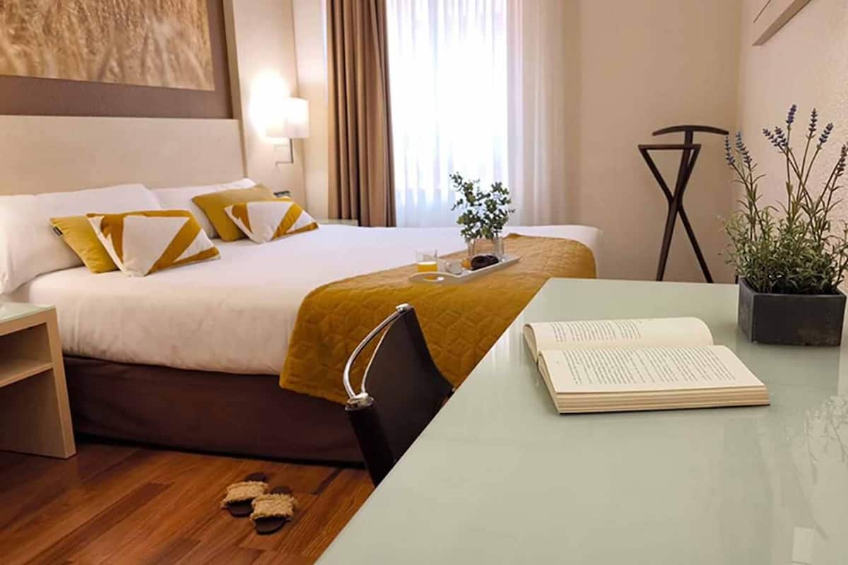 A hotel room with a bed, bedside table, and a desk with a book.
