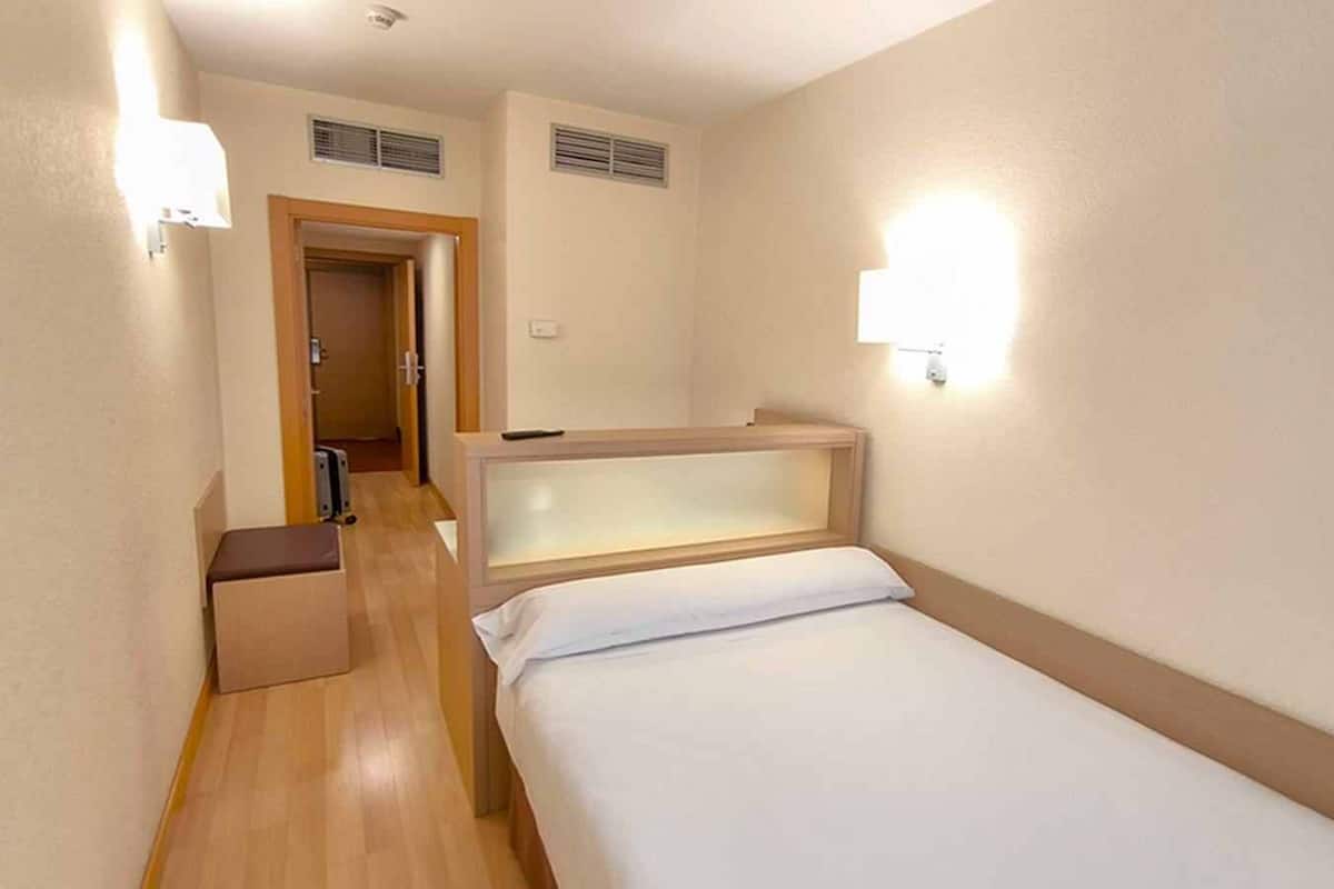 A hotel room with a single bed, a bedside table, a small bench, and two wall-mounted lights.