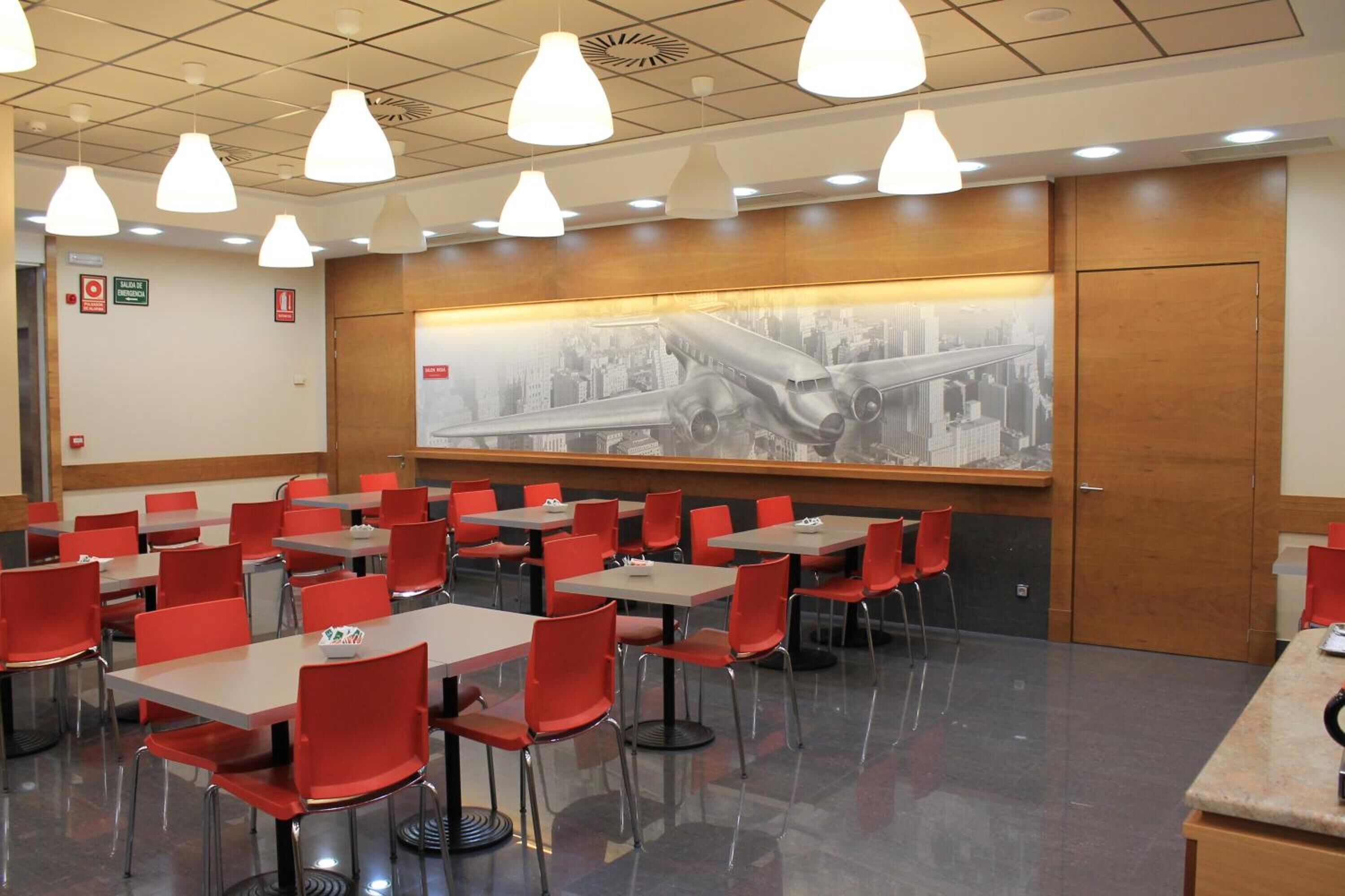 Un espace repas avec des chaises et des tables rouges, une fresque murale représentant un paysage urbain et des luminaires suspendus.