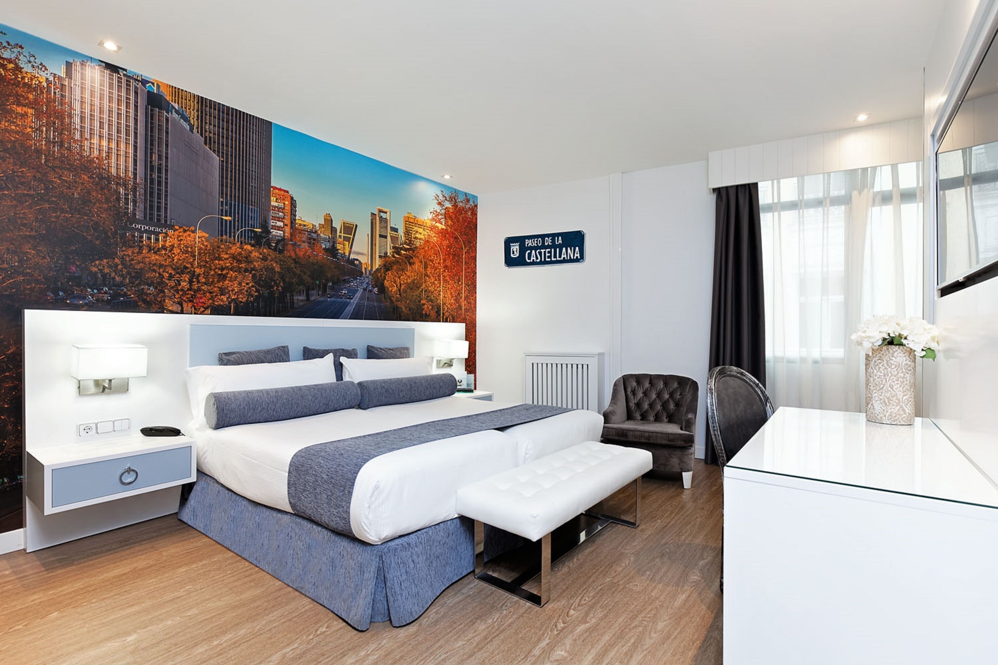 Una habitación de hotel moderna con una cama grande, mesitas de noche, un escritorio, una silla y un mural con vistas a la ciudad.