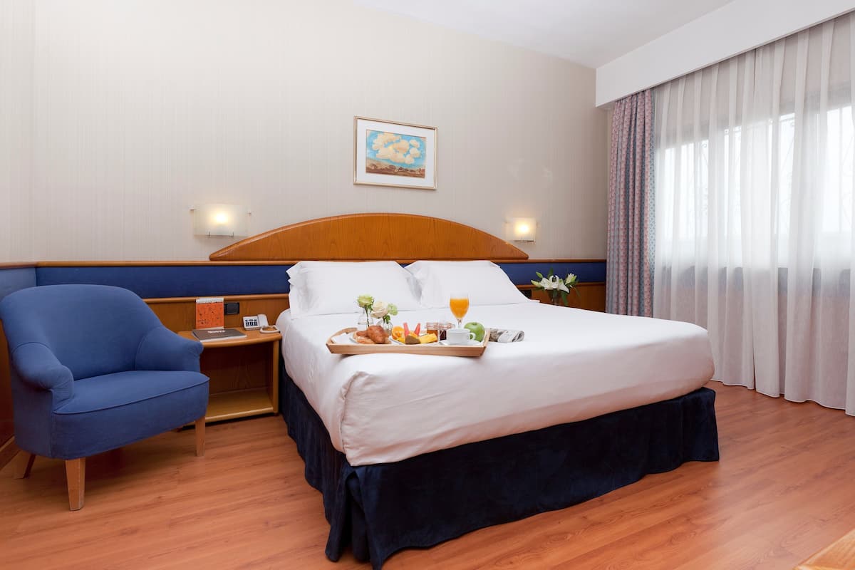 A hotel room with a large bed, a blue armchair, a bedside table with a lamp, and a window with sheer curtains.