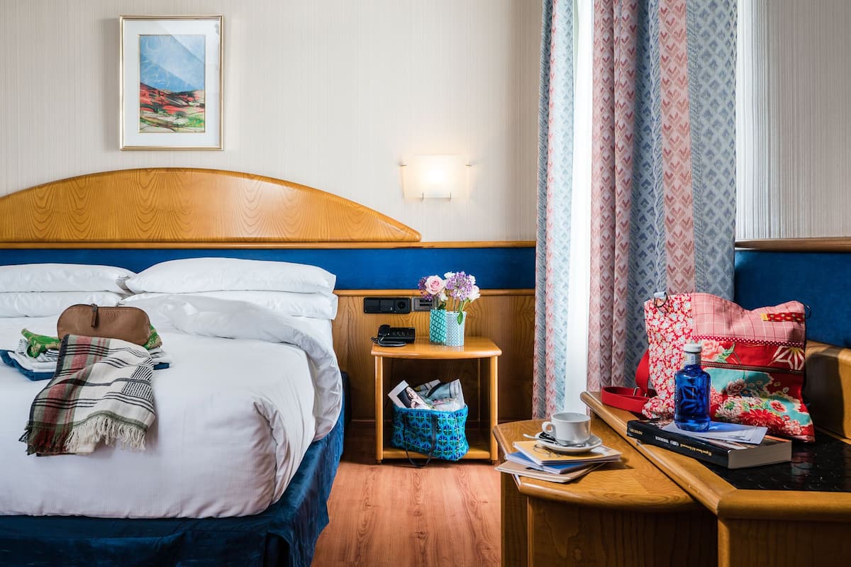 A hotel room with a bed, a bedside table with a lamp, a nightstand with a phone, a cup, a book, and a suitcase.