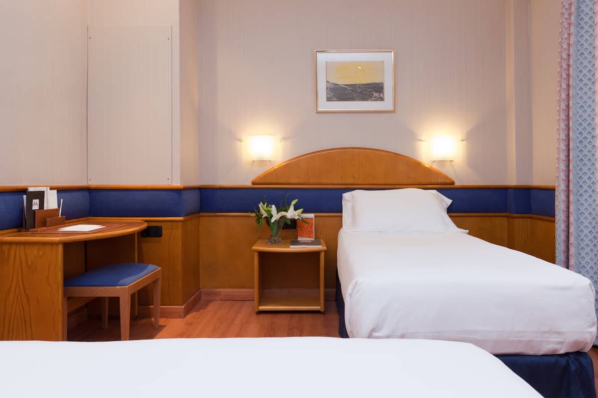 A hotel room with a bed, a desk, a chair, a nightstand with a vase of flowers, and a wall-mounted picture.