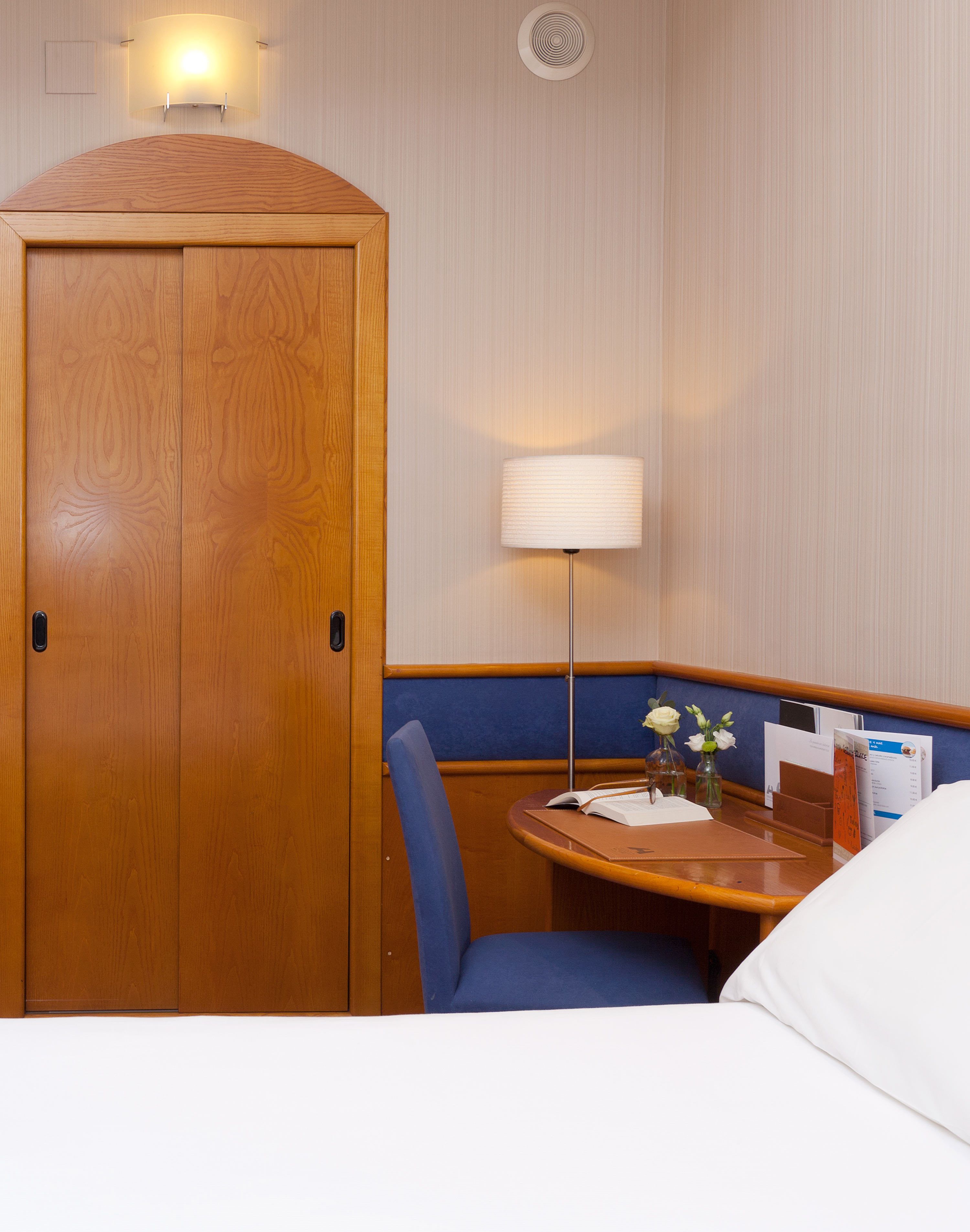 Habitación de hotel con un armario de madera, una mesa redonda con una silla, una lámpara y una mesita con un jarrón de flores.