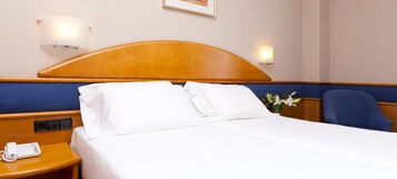 Una habitación de hotel con cabecera de madera, ropa de cama blanca, teléfono en la mesita de noche y una silla azul.