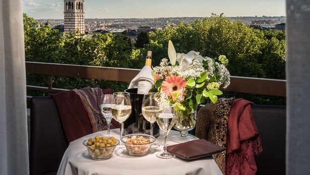 A table set with wine glasses, a bottle of wine, a vase with flowers, and bowls of nuts and fruits, overlooking a cityscape with a prominent tower. - Hotel Agumar Atocha (Madrid)