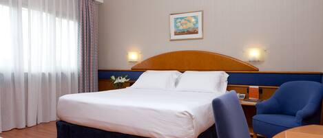 A hotel room with a large bed, a desk with a chair, a nightstand with a lamp, and a painting on the wall.