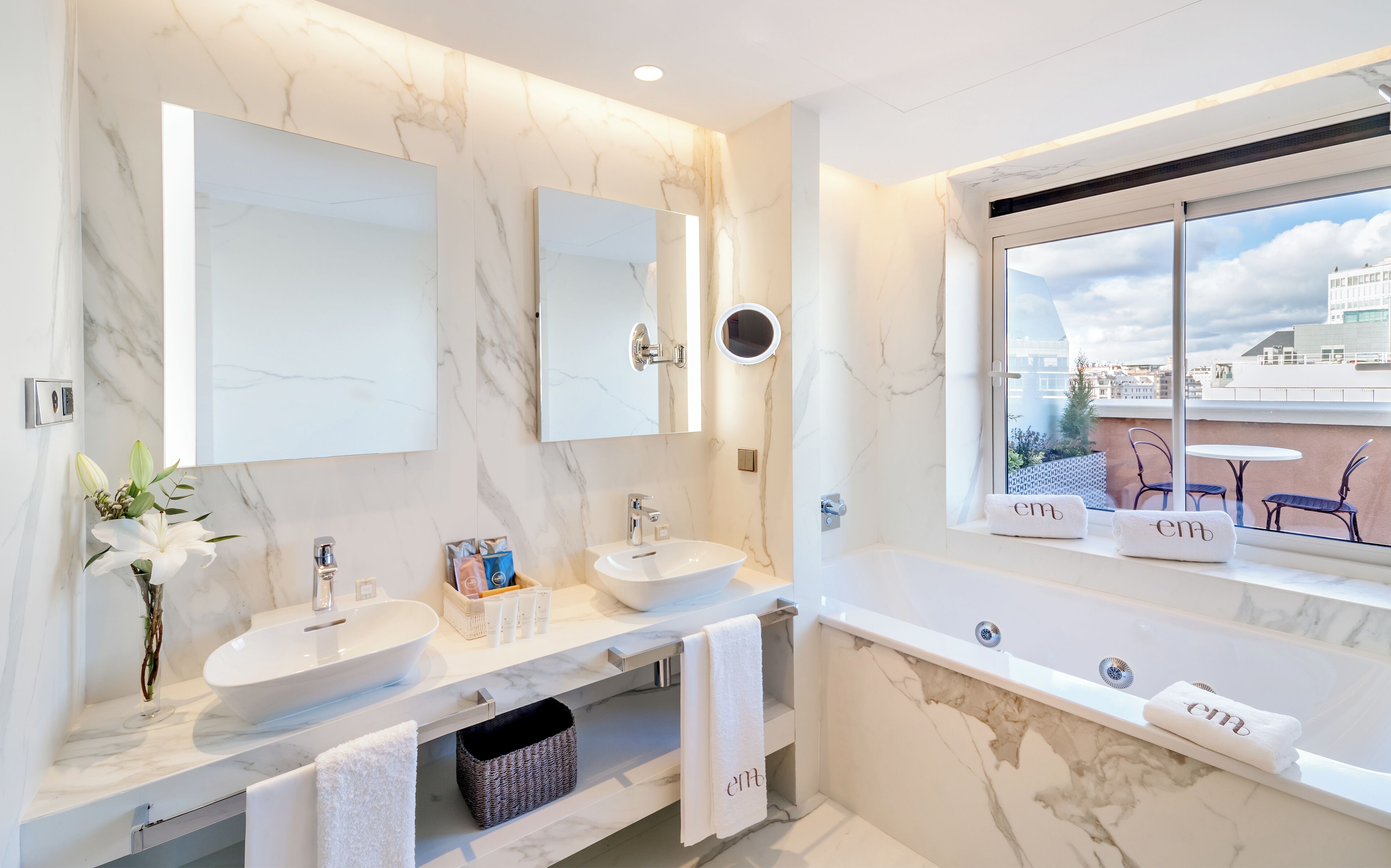 Un baño moderno con un espejo grande, dos lavamanos, una bañera y una ventana con vistas a la ciudad.