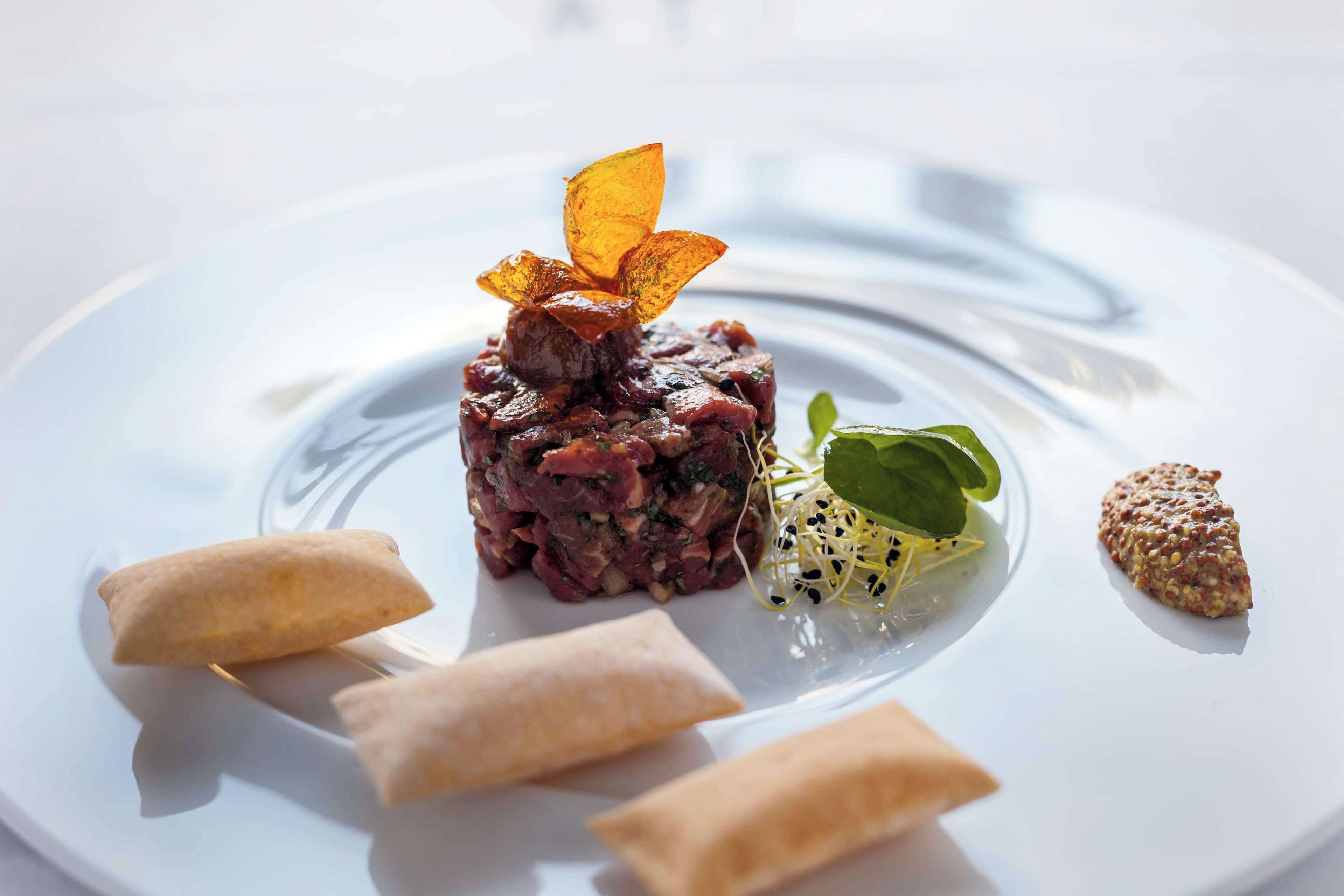 A plate with a dish of minced meat, garnished with a flower-like item, and surrounded by three spring rolls and a small salad.
