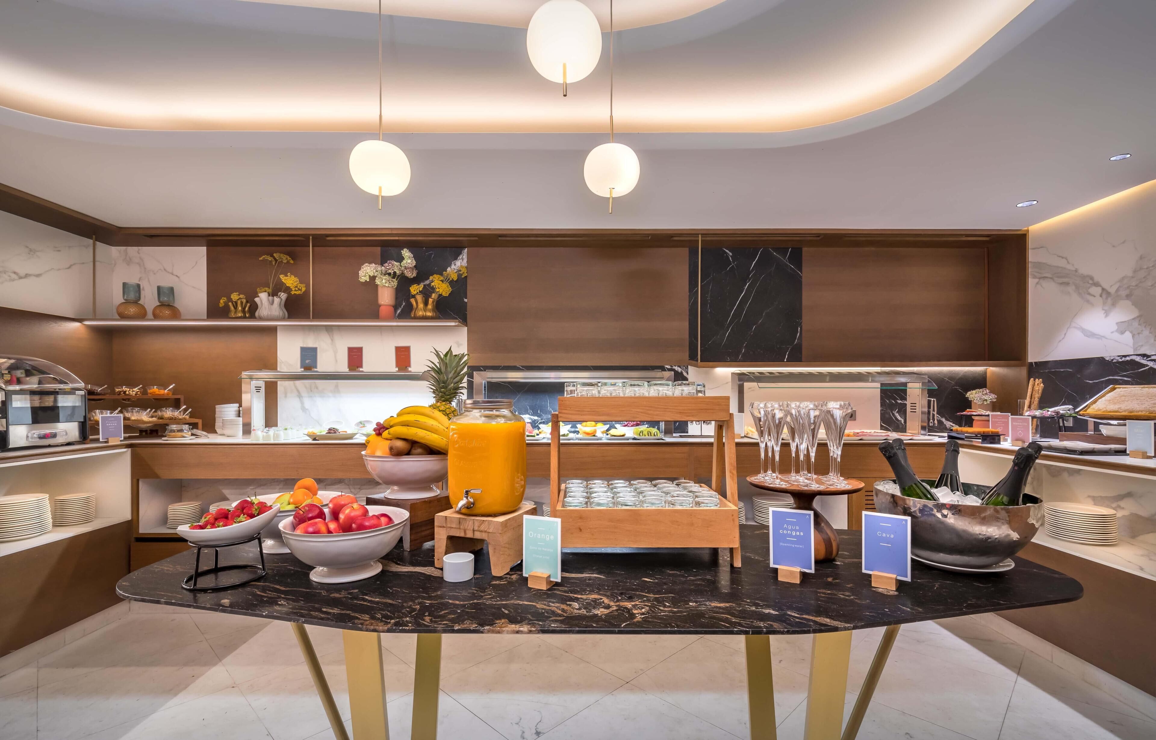 A modern breakfast area with a marble countertop, fruit bowls, a juice dispenser, and a buffet display.