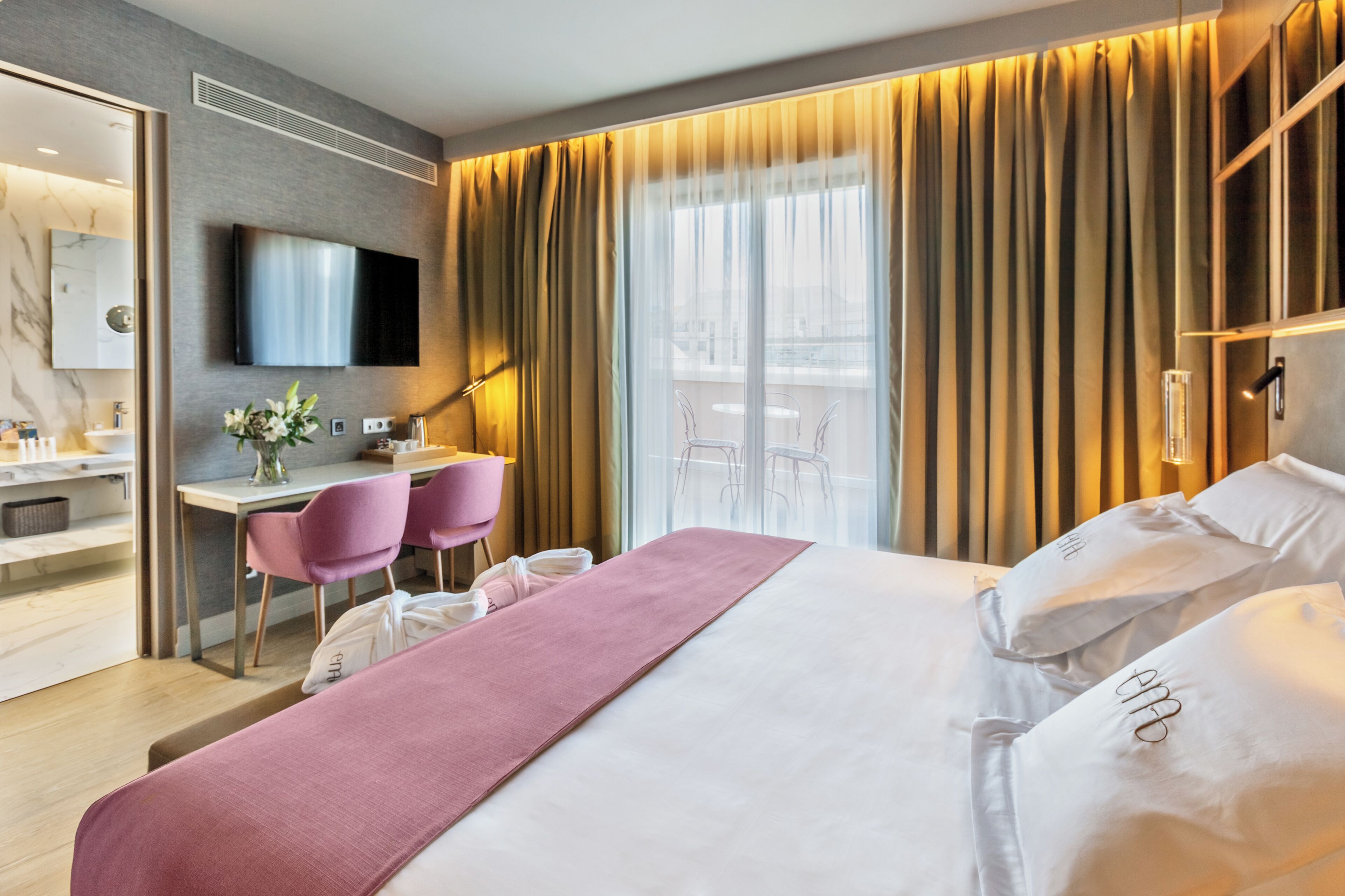 A modern hotel room with a large bed, a desk with a chair, a TV, and a balcony with curtains.
